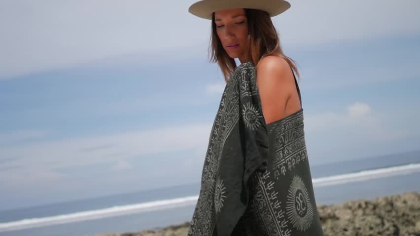 Girl in green bikini with grey hat standing on the beach and wrapping in black towel