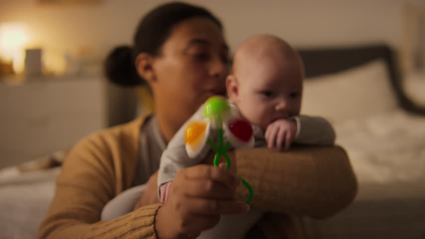 Medium close-up shot of cranky 3-month-old baby in mothers arms, chewing on thumb and sleeve, African American mom shaking rattle, and boy crying, feeling discomfort from teething Royalty-Free Stock Footage #3449196127