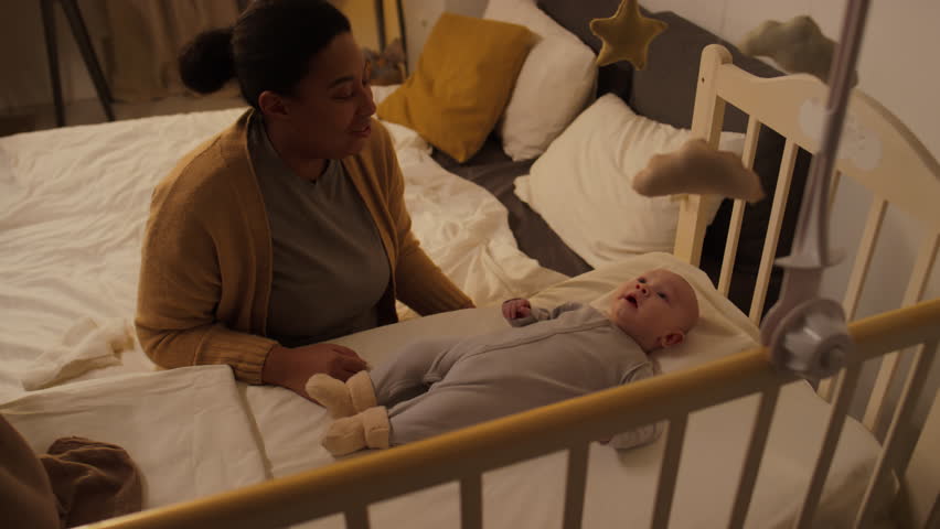 Medium shot of young African American mother sitting in bedroom beside crib, fussy infant boy lying on back, woman talking gently and both looking at baby mobile Royalty-Free Stock Footage #3449222207
