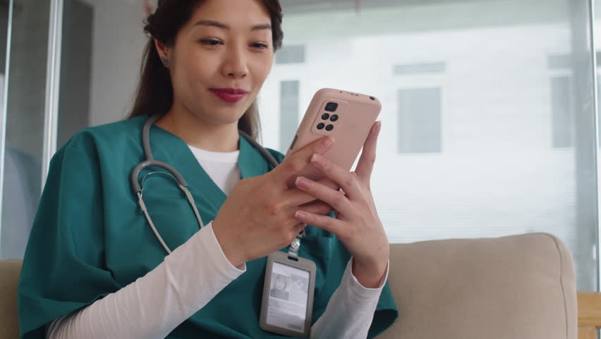 Tilt up of happy Asian nurse in scrubs chatting with friends on smartphone while having break at work