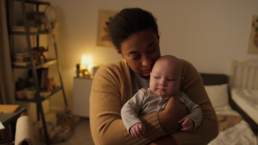 Medium portrait shot of happy young new African American mother standing in bedroom at home, swaying, holding baby son in arms and looking at camera, and boy fussing and crying Royalty-Free Stock Footage #3449228673
