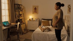 Medium full shot of young Hispanic or African American woman standing in bedroom, doing weight lifting exercise with dumbells to fitness video on laptop, and baby boy lying in cocoon nest on bed