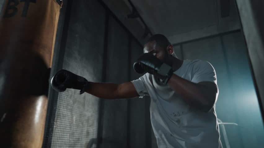 Boxer training hitting punching bag. African american sweated sportsman practicing punches in gym wearing boxing gloves. Workout, martial art, training, sport, boxing practicing, fighting concept.