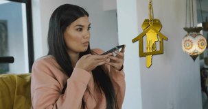Animation of gold house key and key fob over happy biracial woman using smartphone at home. House, home, property, ownership and lifestyle concept digitally generated video.