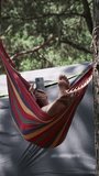 Vertical, Woman using smartphone while swaying in a hammock at summer campsite. Female traveler uses a mobile phone in a pine forest near the tent. Checking social networks, relaxing, lifestyle