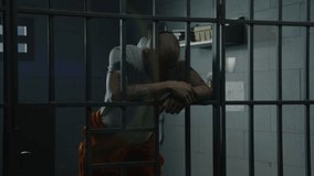Depressed male prisoner in orange uniform stands in prison cell leaning and holding hands on metal bars. Inmate serves imprisonment term for crime in jail. Detention center or correctional facility. - Powered by Shutterstock - Get 15% off with code: PIKWIZARD15