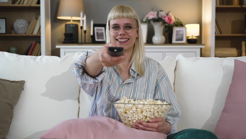 Young happy woman living alone in her new apartment watching television, changing and choosing tv program with remote control and eating popcorn. Leisure weekend activities with movies and tv series Royalty-Free Stock Footage #3449418075