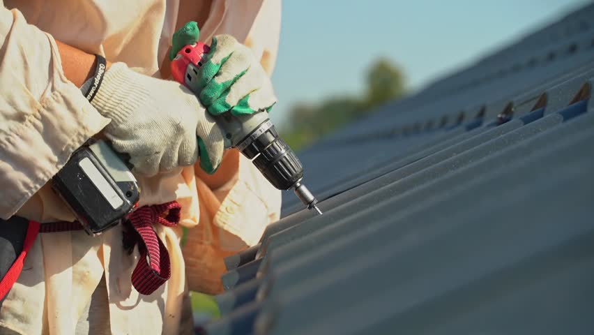 A roofer screws a screwdriver into a black metal tile roof. Roof construction.