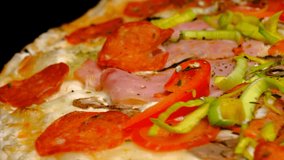 Pizza rotates on a black background, side view, close-up. Pizza with toppings, salami, tomatoes, fresh onions, ham, olives, peppers, cheese. Fast food.