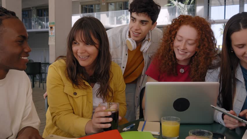 Multiracial generation z students gathered group in campus cafeteria going over syllabus drinking beverages. Joyful young people in community chatting together enjoying break using laptops and tablets