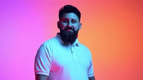 Young bearded man with flirty facial expression looking at camera in neon light against gradient pink-orange studio background. Romantic and love. Concept of beauty, human emotions, facial expression.