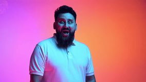 Wow. Bearded man in shock put hands on face and smiling of joy in neon light against gradient pink-orange studio background. Concept of beauty and fashion, human emotions, facial expression. Ad