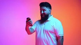 Man dressed casual outfit holds phone and show welcome gesture in neon light against gradient pink-orange studio background. Concept of human emotions, social media, network, connection in distance.
