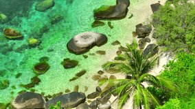 A picturesque coastline adorned with verdant swaying trees, coconut palms standing tall amidst rugged rocks, and azure waters inviting tranquility. Bird's eye view. Gulf of Thailand. 4K.
