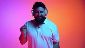 Overjoyed bearded man listening music in headphones and dancing in neon light against vibrant gradient studio background. Concept of relax, human emotions, self-expression, beauty and fashion. Ad