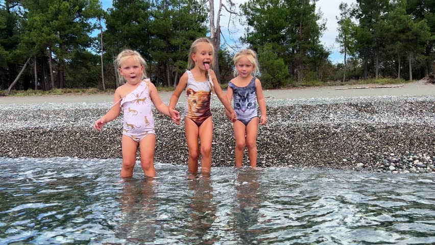 Triplet sisters swim in sea water on a pebble beach, pine trees can be seen behind