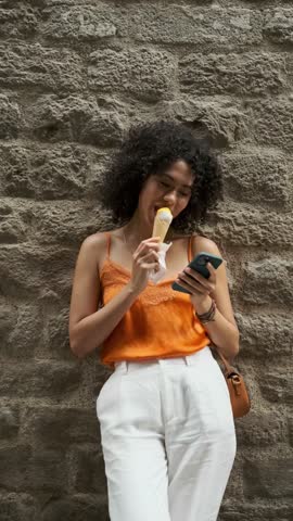 Woman using a mobile phone and taking selfies while eating an ice cream cone on the street.
