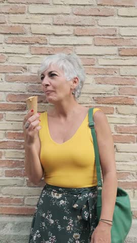 Mature woman enjoying eating an ice cream cone while relaxing outdoors on the street.