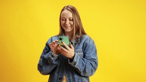 Young woman with freckles in denim jacket using smartphone and speaking by cellular connection against vibrant yellow studio background. Concept of online working, delivery, technology, shopping.
