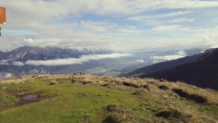 Cinematic aerial: Flying over a mountain top with grassy fields. Valley with a small city (Innsbruck, Tyrol) and steep alpine mountain range (Nordkette) with clouds in the distance. Sunny autumn day