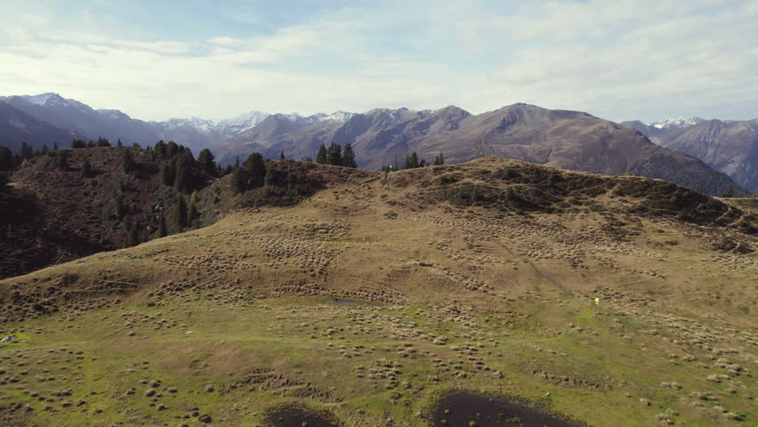 Cinematic aerial: Flying over a mountain top with grassy fields, passing old dead tree on a scenic peak, revealing valley with trees and steep alpine mountain ranges in the distance, sunny autumn day