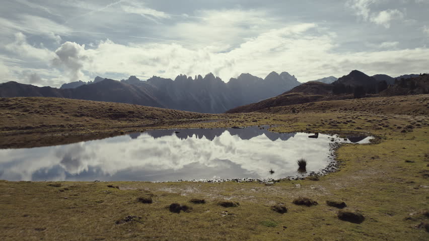 Cinematic aerial: Small lake on a mountain top with grassy fields around. A valley and steep alpine mountain range (Kalkkögel, Tyrol) in the distance reflecting in the water on a sunny autumn day. 4K