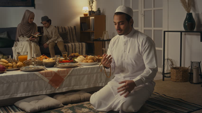 Side footage of muslim Arab in white thawb and cap performing takbir with beads sitting on floor near abundant festive dinner table on Eid al-Fitr with his wife and son reading Koran in background Royalty-Free Stock Footage #3449733499