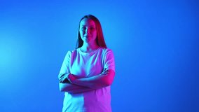 Young woman in white T-shirt looking at camera with smiling and crossed hands in neon light against blue gradient background. Concept of beauty, fashion, self-expression, human emotions.