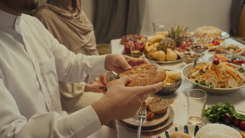 Side tilt footage of smiling bearded muslim man sharing flatbread or pita with family members during festive dinner celebrating Eid al-Fitr at home Royalty-Free Stock Footage #3449749115