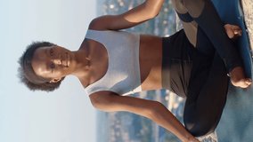 Vertical video of African American woman wearing sportswear during meditation. Young female comfortably sitting in lotus position on fitness mat. Yoga concept. Well-being. Soul and body.