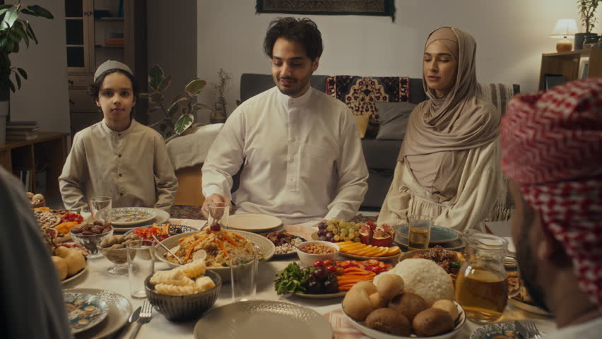 Over shoulder footage of unidentified muslim man passing plate with potatoes and flatbread to positive family members during gathering at holiday table on Eid al-Fitr at home Royalty-Free Stock Footage #3449781065