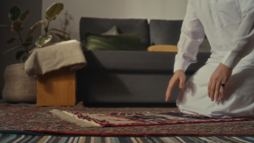 Side ground level footage of muslim man in cap and thawb prostrating while praying to Allah on prayer map at home Royalty-Free Stock Footage #3449797765