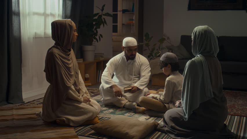 Full footage of father in white muslim cap and kandora explaining basic rules of islam with Koran in hand to little son. Sister and mother sitting nearby in hijabs and listening Royalty-Free Stock Footage #3449819901