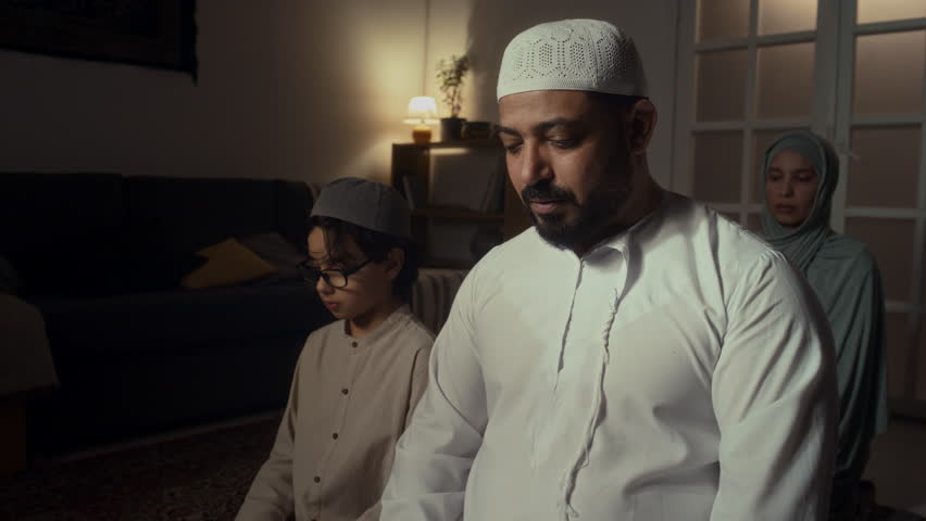 Side footage of Biracial man in muslim cap and thawb praying to Allah together with his son and wife who sitting behind them during namaz at home Royalty-Free Stock Footage #3449819989