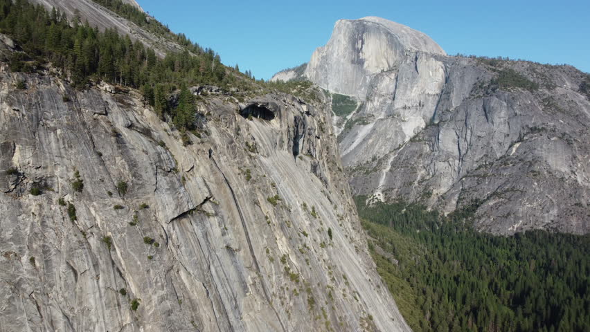 Yosemite National Park, the aerial shot going backward. The shot shows the half dome and a clear view of the granite monoliths on the closest mountain. The forest is under the camera