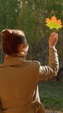 A red-haired girl holds an autumn leaf in her hand and covers the sun with it. The wind is tearing yellow leaves from the trees. Autumn has come. Vertical video for shorts.