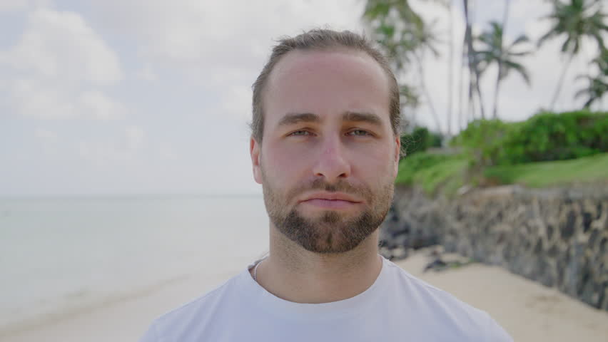 Nice unshaved man in white T-shirt looking at camera, opening mouth wide, showing great fear, white ocean and sky with green palm trees on the background. High quality 4k footage Royalty-Free Stock Footage #3450013881