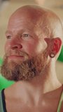 Vertical Portrait of Strong Masculine Male Athlete Smiling and Posing at Rock Climbing Gym with Bouldering Wall Background. Handsome Happy Bald Man with Ginger Beard Wearing Colorful Undershirt