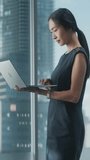 Vertical Portrait: Successful Businesswoman in Stylish Dress Working on Laptop, Standing Next to Window in Big City. Confident Female CEO Analyze Financial Projects. Manager at Work Planning Campaign.