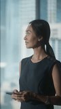 Vertical Screen: Businesswoman in Black Dress Walking in Modern Office, Using Smartphone, Looking out of the Window on Big City with Skyscrapers. Confident Female CEO Working on Financial Projects.