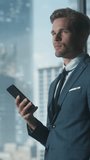 Vertical Screen: Confident Businessman in a Suit Standing in Modern Office, Using Smartphone, Looking out of the Window on Big City with Skyscrapers. Successful Finance Manager Planning Work Projects.