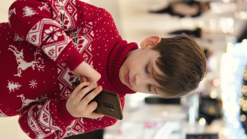 A boy in a New Year's sweater happily communicates on a smartphone in a shopping center. Caucasian boy 9 years old in a shopping center with a smartphone and a festive sweater. vertical video Royalty-Free Stock Footage #3450153041