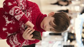 A boy in a New Year's sweater happily communicates on a smartphone in a shopping center. Caucasian boy 9 years old in a shopping center with a smartphone and a festive sweater. vertical video
