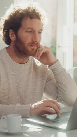 Vertical Screen: Adult Man with Ginger Curly Hair Using Laptop Computer, Sitting in Living Room in Apartment. Joyful Man is Working from Home, Online Shopping, Watching Videos or Writing Emails. Royalty-Free Stock Footage #3450172719