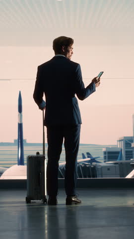 Vertical Screen. Airport Terminal: Businessman with Rolling Suitcase Walks, Uses Smartphone App for e-Business. Back View Silhouette of Traveling Man Waits for Flight in Boarding Lounge of Airline Hub
