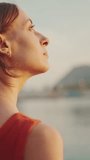 VERTICAL VIDEO: Close-up, girl stands on the seashore and looks at the bay and fixes her hair. Closeup portrait of a young woman looks at the sea in the morning time.