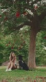 VERTICAL VIDEO: Cute girl with black dog is resting under tree on the lawn. Happy dog rejoices being on walk in the outdoor air. Animal training outdoors