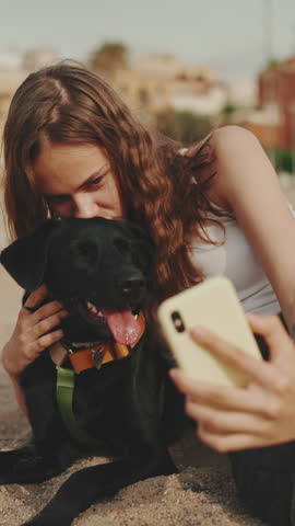 VERTICAL VIDEO: Clouse-up, cute girl with long wavy hair wearing white top takes selfie while sitting on the sand with black dog on modern building background. Girl hugging her pet