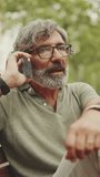 VERTICAL VIDEO: Friendly middle-aged man with gray hair and beard wearing casual clothes sits on bench and uses mobile phone. Mature gentleman in eyeglasses talking on cellphone