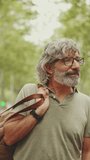 VERTICAL VIDEO: Middle-aged man with gray hair and beard walks and looks around. Mature gentleman in eyeglasses with bag on his shoulder walks through the square on the cityscape background